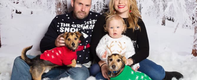 a family wear christmas jumpers and sit on the ground in a snowy christmas photo shoot set