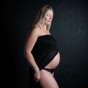 A pregnant lady is photographed in a black dress that opens to reveal her bump against a black background at a professional pregnancy photo shoot