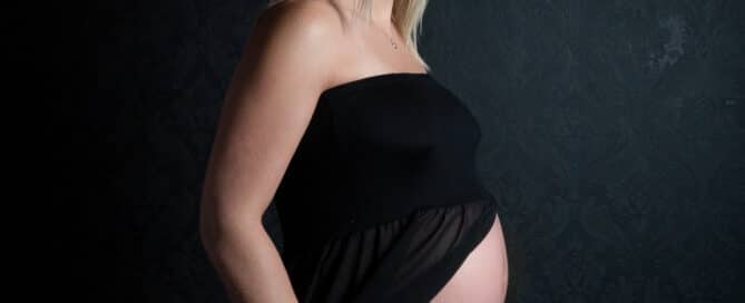 A pregnant lady is photographed in a black dress that opens to reveal her bump against a black background at a professional pregnancy photo shoot