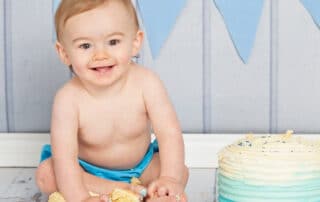 a little boy smiles while he grabs handfuls of cake at his first birthday photo shoot