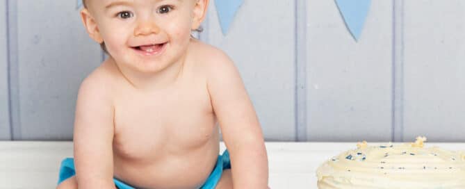 a little boy smiles while he grabs handfuls of cake at his first birthday photo shoot