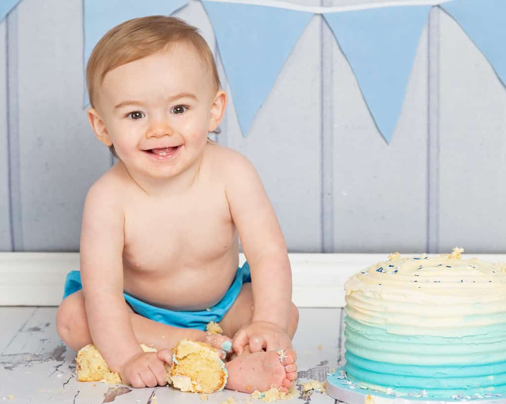 a little boy smiles while he grabs handfuls of cake at his first birthday photo shoot