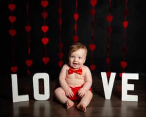 a little boy poses in a red bowtie in front of a curtain of hearts in a Love themed professional photo