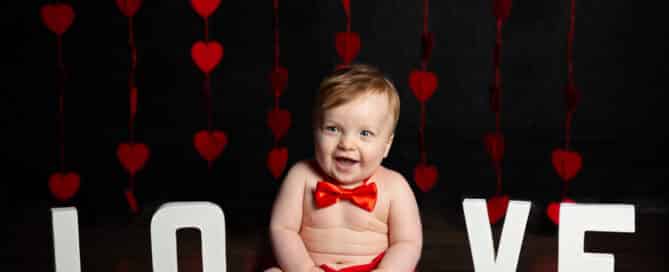a little boy poses in a red bowtie in front of a curtain of hearts in a Love themed professional photo