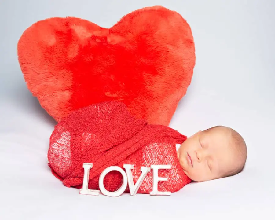 a sleeping newborn in a red wrap in front of a red heart cushion