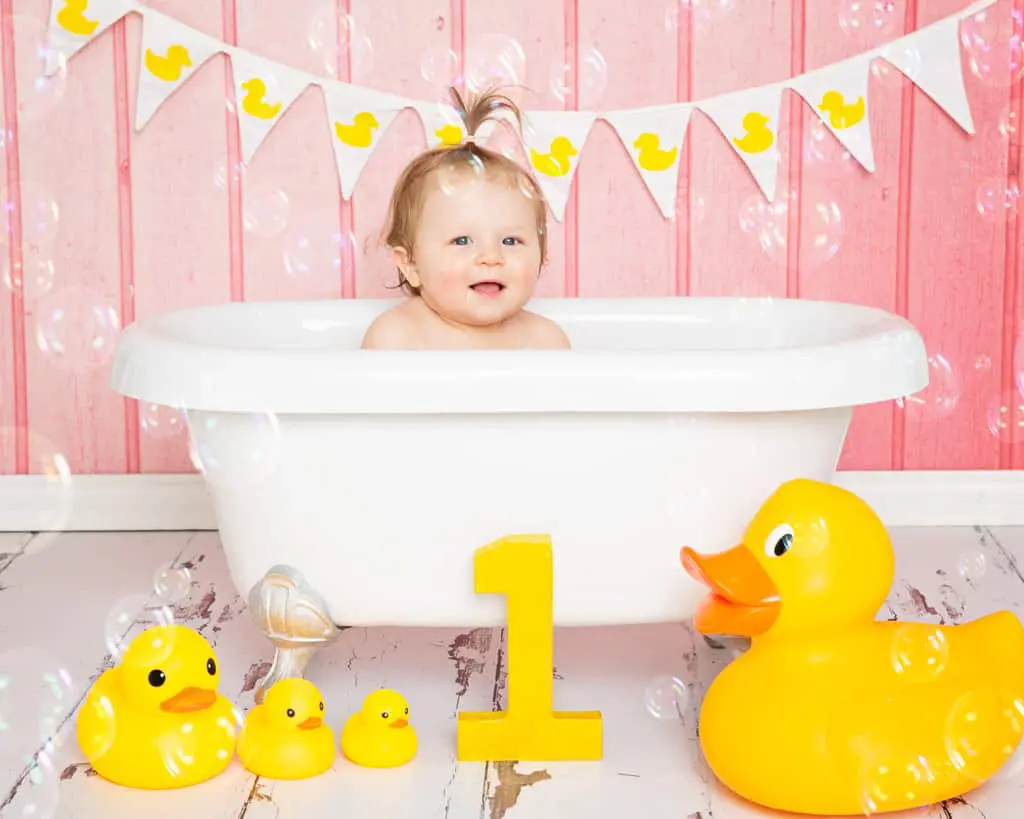 Cake Smash Photography East Grinstead West Sussex A little girl sits in the bathtub against a pink background at her first birthday tub splash photo shoot