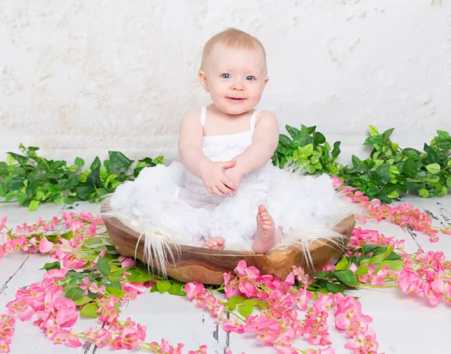 vBaby Photography East Grinstead West Sussex a little girl sits in a bowl surrounded by flowers in a white pettidress