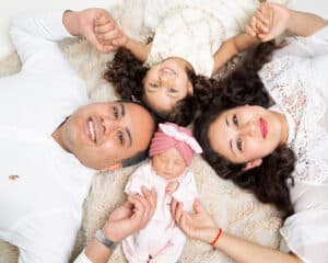A family of four, including a sleeping newborn, lie on the floor and look up at the camera