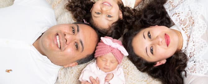 A family of four, including a sleeping newborn, lie on the floor and look up at the camera