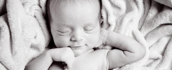 A black and white image of a sleeping newborn on their back with one hand behind their head looking very relaxed