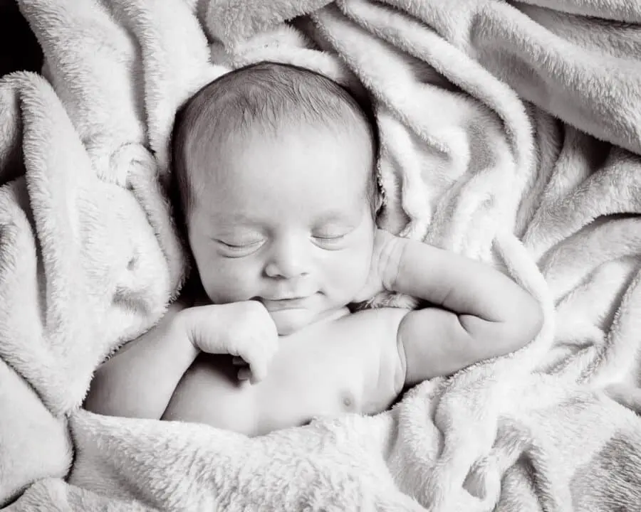A black and white image of a sleeping newborn on their back with one hand behind their head looking very relaxed
