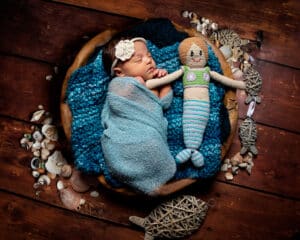 A sleeping newborn in a wooden bowl with a mermaid crocheted toy surrounded by shells