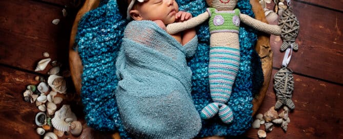 A sleeping newborn in a wooden bowl with a mermaid crocheted toy surrounded by shells