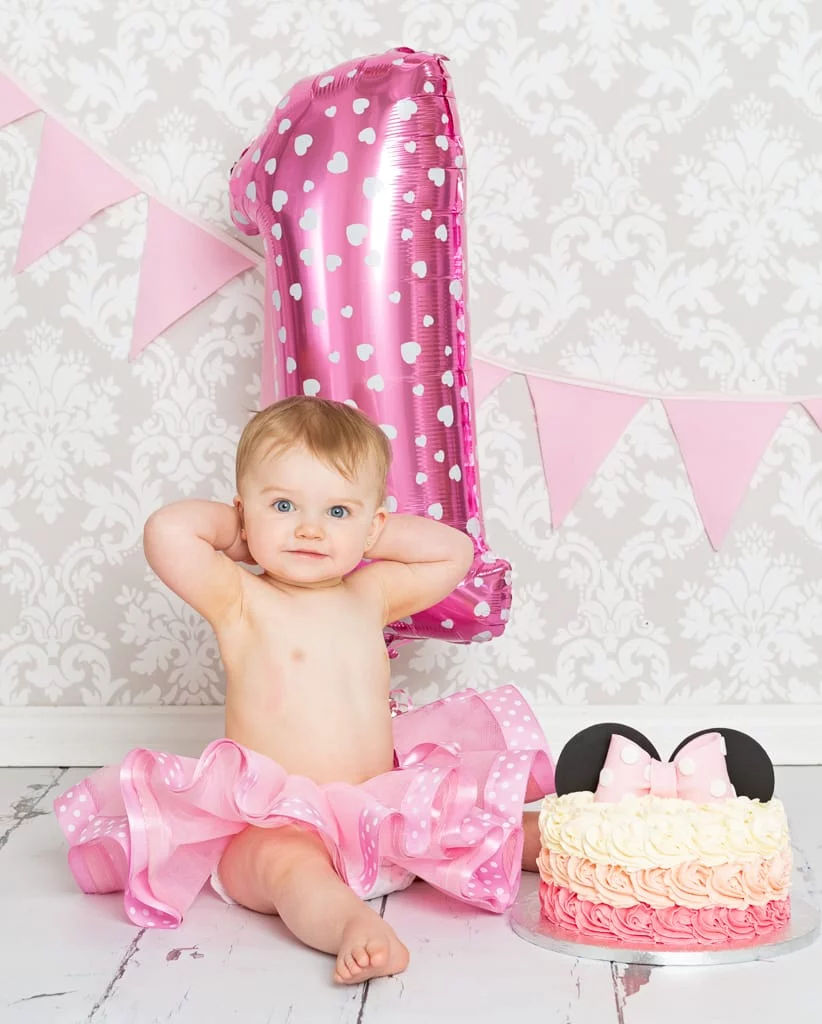 a little girl sits with a balloon and a Minnie Mouse themed cake smash cake