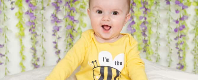 a little baby in a bee happy vest poses against a flower wall