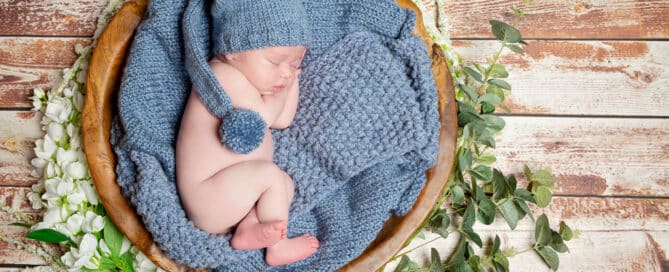 A little newborn boy asleep in a wooden bowl surrounded by flowers and lying on a blue blanket