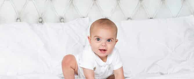 a baby in a white vest moves to crawl on a white duvet covered bed