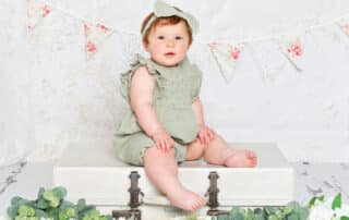 a baby girl in a sage green romper sat on a vintage suitcase