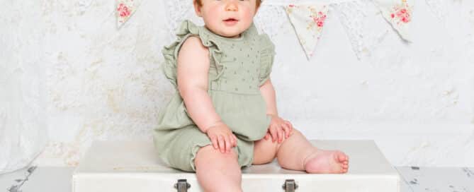 a baby girl in a sage green romper sat on a vintage suitcase