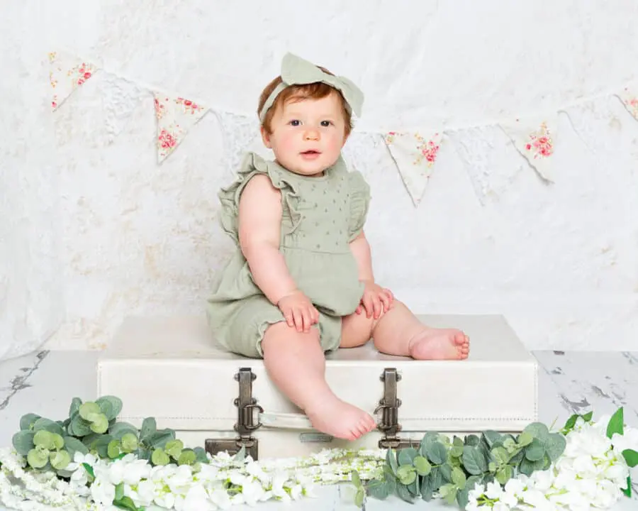 a baby girl in a sage green romper sat on a vintage suitcase