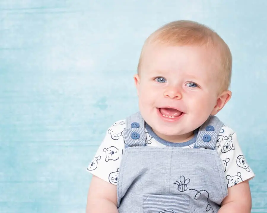 a blue eyed baby boy smiles for the camera against a matching blue background