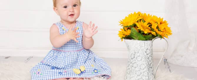 a baby girl in a blue gingham dress next to a vase of sunflowers
