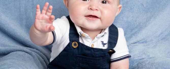 a baby in blue dungarees sits on a cushion and waves at the camera