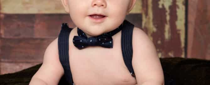 little boy in a black bow tie sits on a cushion against a dark wood background