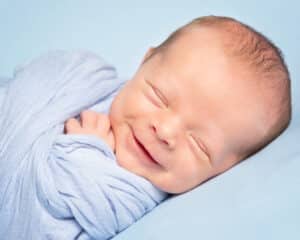 A smiling newborn wrapped in blue and lying on a blue blanket asleep