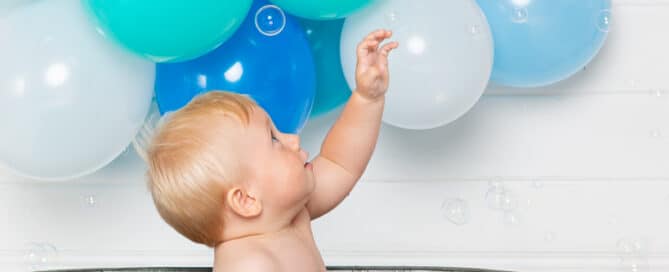 a little boy reaches up to play with some bubbles in the air as he sits in the bathtub in front of a bunch of balloons at his first birthday photo shoot