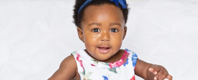 a little girl in a romper sitting on a white bed set