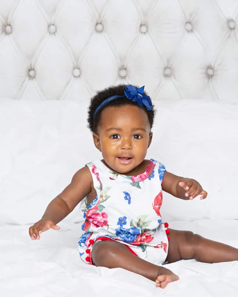 a little girl in a romper sitting on a white bed set