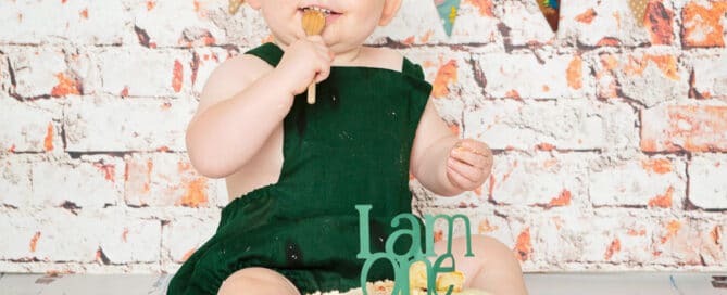 a little boy in a gren romper eats cake at his first birthday shoot