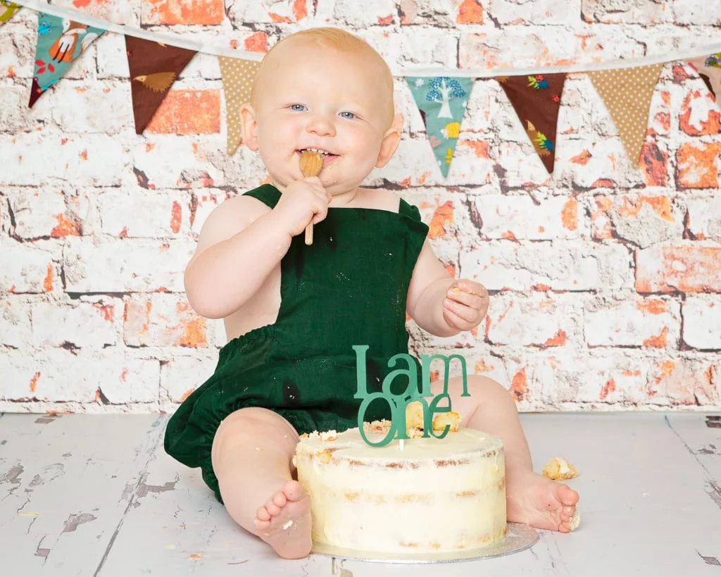 a little boy in a gren romper eats cake at his first birthday shoot