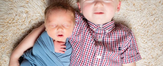 A wrapped newborn lying on the floor with his big brother who is looking up at the camera