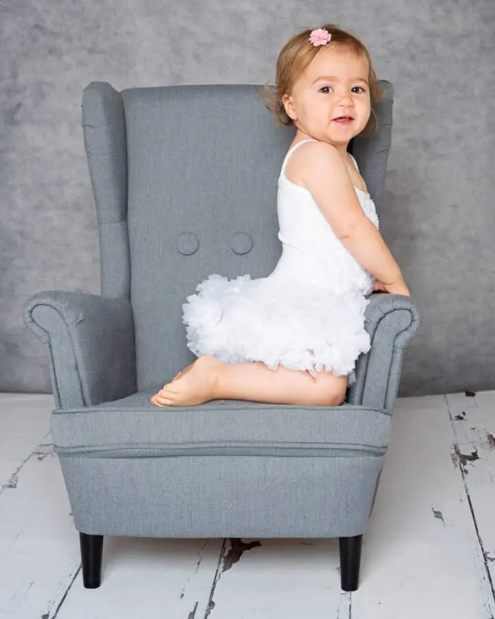 a little girl on a mini wingback chair in a white dress at a photo shoot