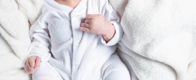 a litlte boy in a white baby sleepsuit against a white blanket backdrop