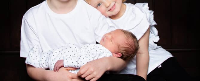 an older brother and sister wear matching white tshirts and hold their newborn sibling