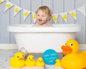 Cake Smash Photography East Grinstead West Sussex a little boy with blond curly hair sits in a white bathtub with bright yellow ducks at his first birthday photo shoot