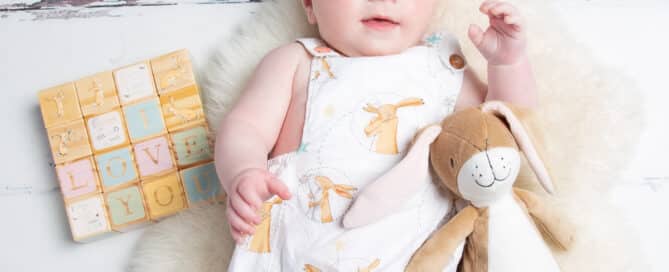 a baby poses in a Nutbrown Hare romper with a stuffed toy