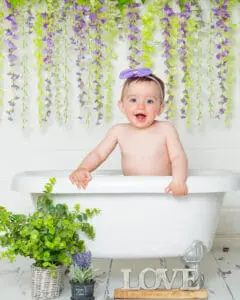  alittle girl is sitting in a white miniature roll top bath against a backdrop of a flower wall at a professional first birthday photo shoot