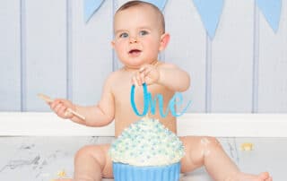 a baby sits behind a giant blue cupcake against a blue background at a first birthday photoshoot