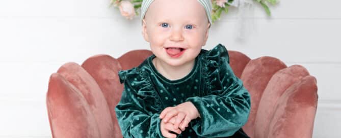 a little girl in a green dress on a pink chair in front of a flower hoop