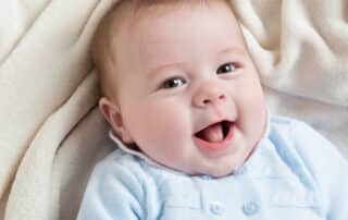 a little boy is lying on a cream blanket, looking up at the camera and smiling