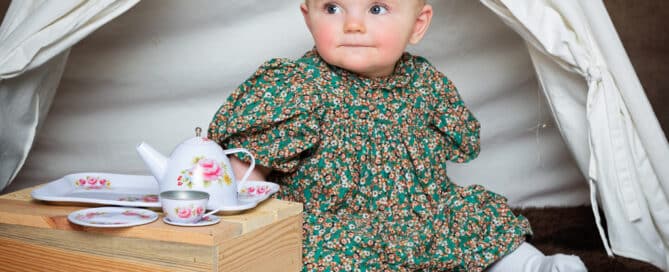 A little girl in a floral dress sits with tea time props in a teepee