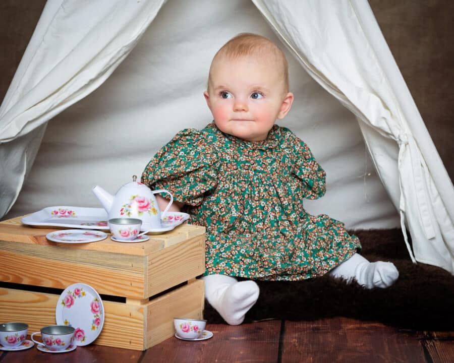 A little girl in a floral dress sits with tea time props in a teepee
