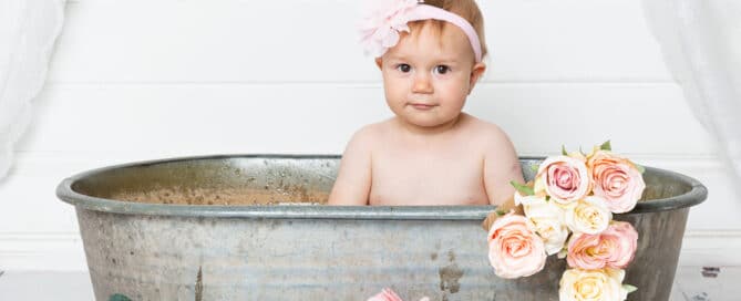 a little girl sits in a tin tub surrounded by peach flowers at her first birthday photo shoot