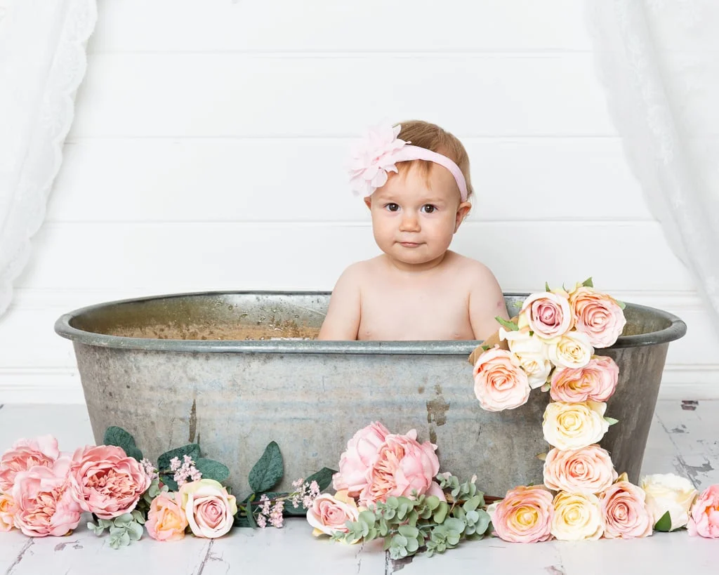 a little girl sits in a tin tub surrounded by peach flowers at her first birthday photo shoot