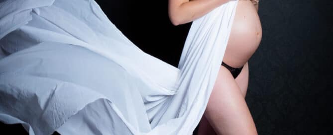 A woman in a flowing white dress poses against a black background for a professional maternity photo in my studio in East Grinstead