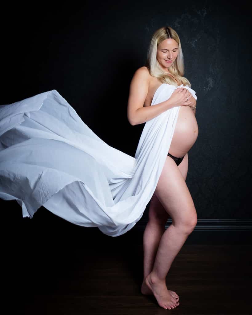 A woman in a flowing white dress poses against a black background for a professional maternity photo in my studio in East Grinstead